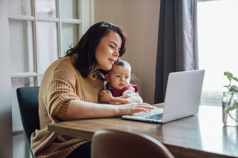Mom holding baby and working from home on laptop at dining room