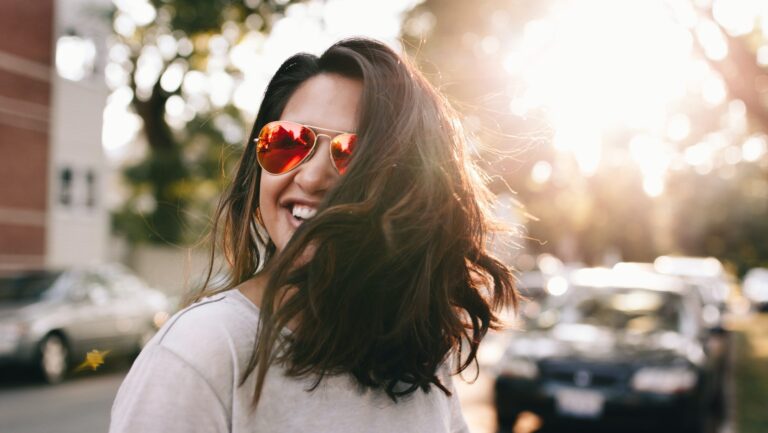 Woman with shoulder-length brown hair wearing a t-shirt and red-lensed sunglasses smiles at the camera. The sun shines through the trees in the background.