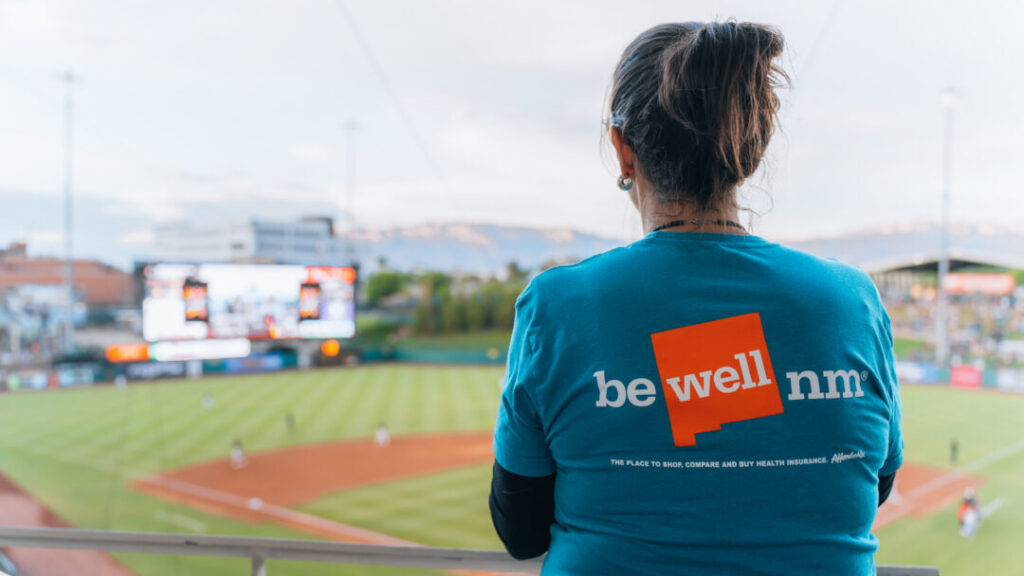 Woman in a blue shirt with the BeWell logo on the back looking out onto the Isotopes baseball field.