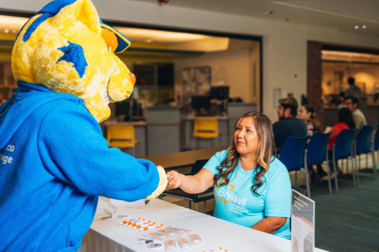 Woman sitting down in BeWell shirt shaking the hand of CNM mascot the Suncat